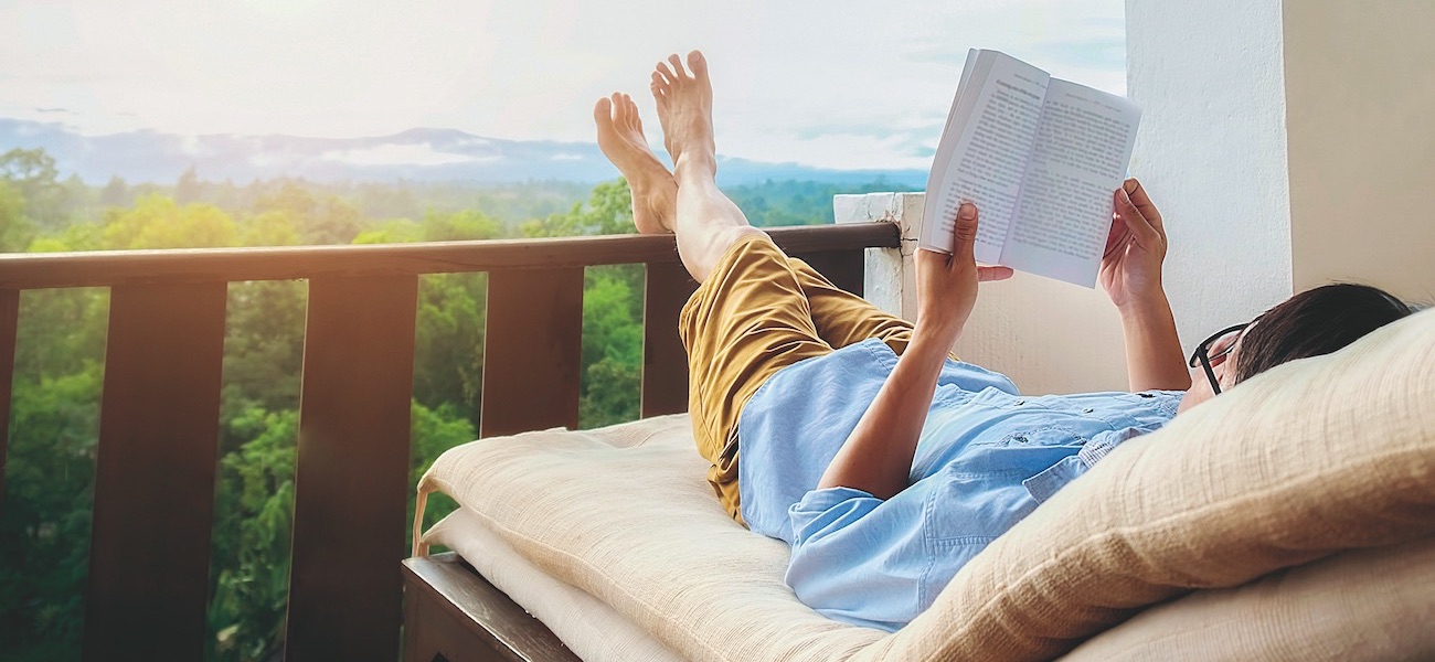 Rear view of asian man relaxing on a sofa and holding book on bed at home terrace with beautiful green background view. Relaxing concept.